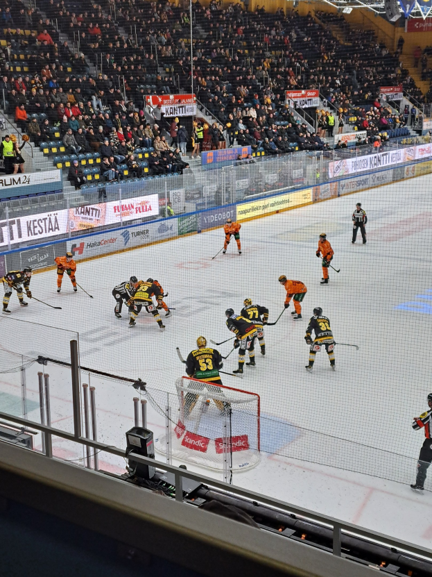 Eine Gruppe von Menschen, die Hockey auf einem Eisstadion spielen, umgeben von einem Zaun, mit Zuschauern im Hintergrund und Bannern unter Stadionbeleuchtung.