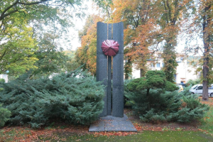 Große Metallskulptur in einem Park mit Bäumen, Pflanzen und zerrissenen Blättern, umgeben von einer Straße mit Fahrzeugen, Gebäuden im Hintergrund und einem klaren blauen Himmel.