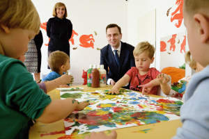 Kinder beim Malen an einem Tisch mit Farbe an den Händen, Papieren und Flaschen, beaufsichtigt von einer lächelnden Frau, mit Kunstwerken an der Wand im Hintergrund.