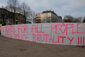 Eine Gruppe von Menschen, die auf dem Boden stehen und ein Banner halten, auf dem "Rechte für alle Menschen - Stoppt Polizeigewalt" steht, mit einem Straßenschild, einem Schild, Bäumen, Gebäuden mit Fenstern und einem bewölkten Himmel im Hintergrund.