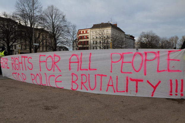 Eine Gruppe von Menschen, die auf dem Boden stehen und ein Banner halten, auf dem "Rechte für alle Menschen - Stoppt Polizeigewalt" steht, mit einem Straßenschild, einem Schild, Bäumen, Gebäuden mit Fenstern und einem bewölkten Himmel im Hintergrund.