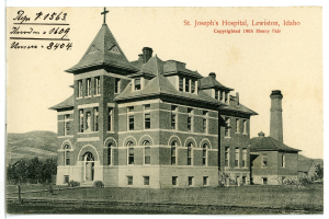 Ein Schwarz-Weiß-Foto des St. Joseph-Krankenhauses in Lewiston, Idaho, das ein großes Gebäude mit Fenstern, Treppen mit Geländern, einen Schornstein auf der rechten Seite und Text oben zeigt, mit Bäumen, Hügeln und Himmel im Hintergrund.