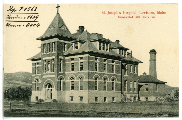 Ein Schwarz-Weiß-Foto des St. Joseph-Krankenhauses in Lewiston, Idaho, das ein großes Gebäude mit Fenstern, Treppen mit Geländern, einen Schornstein auf der rechten Seite und Text oben zeigt, mit Bäumen, Hügeln und Himmel im Hintergrund.