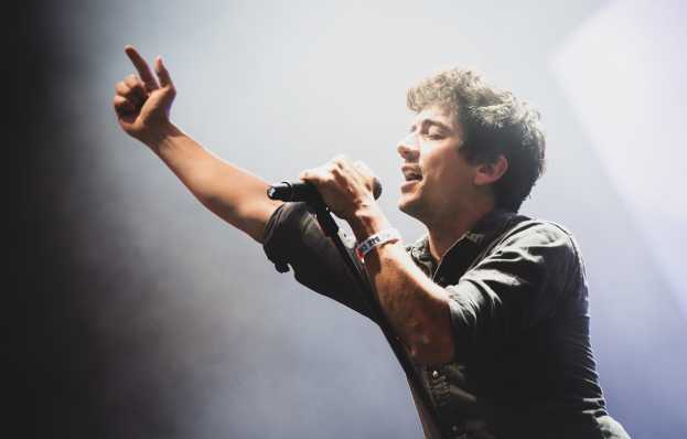 Ein Mann in schwarzem Hemd und Jeans singt leidenschaftlich in ein Mikrofon auf einer hell erleuchteten Bühne, während das Publikum im Hintergrund klatscht und jubelt.