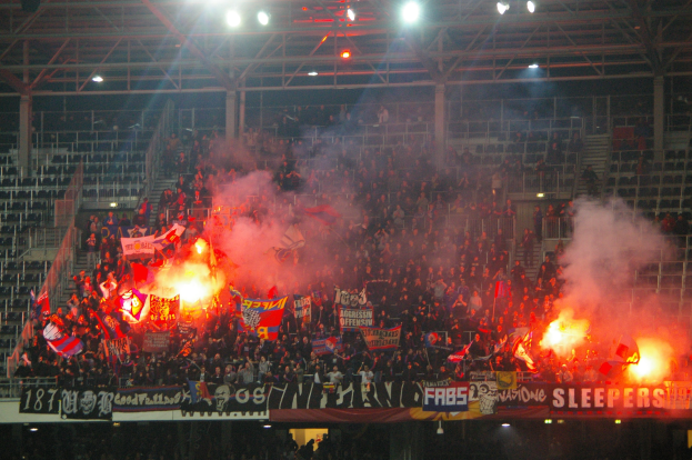 Eine große Menge Menschen in einem Stadion hält Fahnen und Banner mit Feuerzeugen und Rauch drumherum, unter einer Decke mit Deckenleuchten.