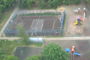 Eine Luftaufnahme eines Parks mit einem eingezäunten Basketballplatz, Straßenlaternen, Bäumen, Gras, Rutschen und anderen Spielplatzgeräten.