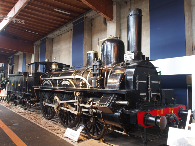 Eine große, alte Dampflokomotive in einem Museums-Setting umgeben von Eisenbahnschienen, Informations-Tafeln und anderen Ausstellungsstücken, mit beleuchteter Decke und Wand im Hintergrund.