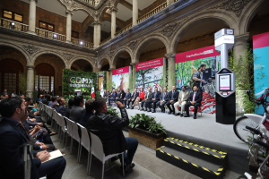 Eine Gruppe von Menschen sitzt in Stühlen und schaut auf einen Mann auf einer Bühne, der ein Mikrofon hält, während eine Pressekonferenz für den CDMX-Nachhaltigkeitsenergiegipfel stattfindet, mit Fahrrädern auf der rechten Seite und verschiedenen architektonischen Elementen im Hintergrund.