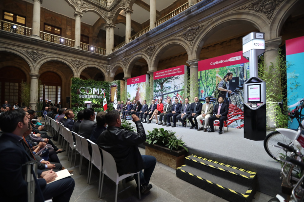 Eine Gruppe von Menschen sitzt in Stühlen und schaut auf einen Mann auf einer Bühne, der ein Mikrofon hält, während eine Pressekonferenz für den CDMX-Nachhaltigkeitsenergiegipfel stattfindet, mit Fahrrädern auf der rechten Seite und verschiedenen architektonischen Elementen im Hintergrund.