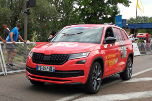 Ein rotes Auto fährt auf einer Straße neben einer Menschenmenge, einige tragen Mützen, mit Maschendraht auf der linken Seite, einem Verkehrsampelmast, Bäumen, einer Brücke, einem Fahnenmast und einem bewölkten Himmel im Hintergrund.