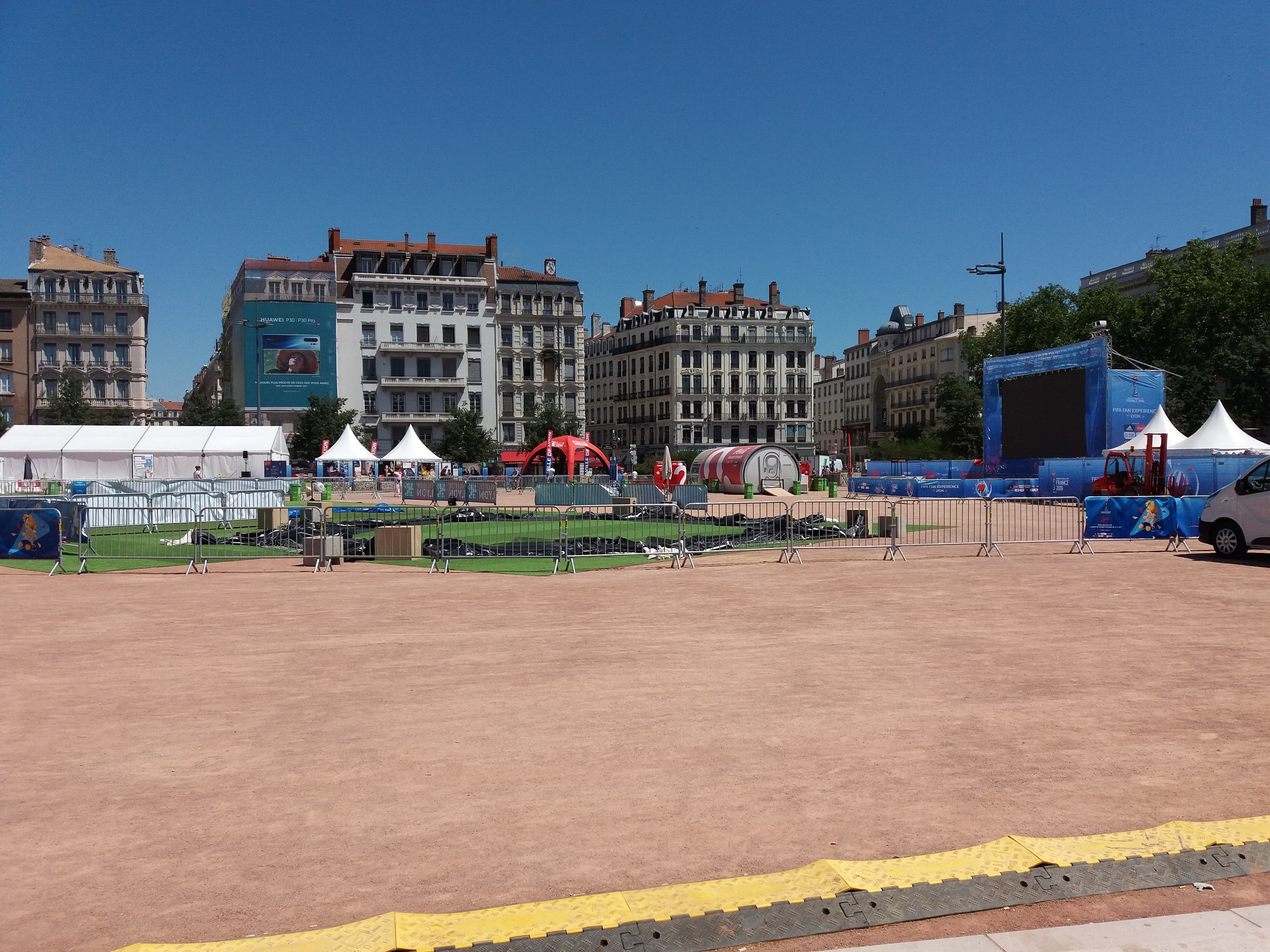Großer städtischer Platz voller Zelte, umgeben von Gebäuden, Bäumen und Fahrzeugen unter einem klaren blauen Himmel.