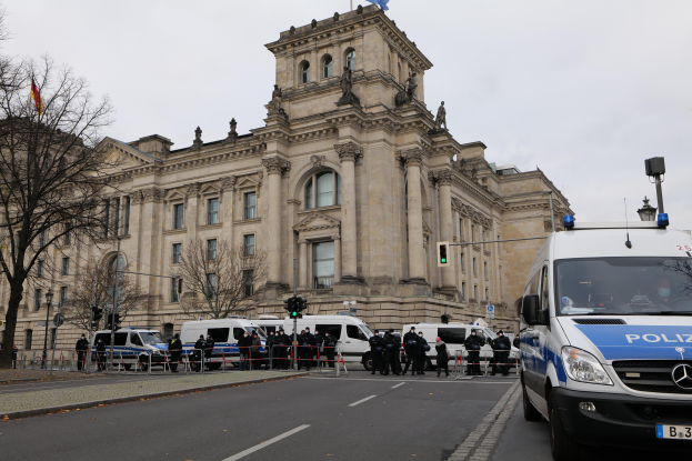 Eine Gruppe von Polizisten steht vor dem Reichstaggebäude in Berlin, Deutschland, mit Fahrzeugen, einem Zaun, Verkehrsampeln, Laternen, Bäumen und Flaggen im Hintergrund, unter einem klaren Himmel.