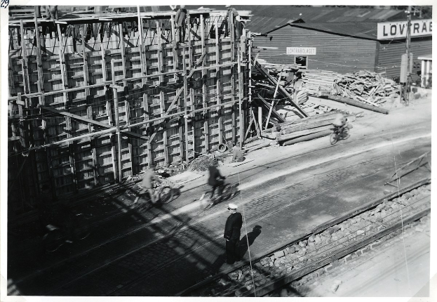 Schwarz-weiß-Foto einer Baustelle mit einem teilweise errichteten Gebäude, Menschen auf Fahrrädern und zu Fuß, Bahnschienen, Pfosten, Bretter und Text unten.