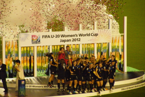 Gruppe von Frauenfußballspielerinnen in schwarzer Uniform auf einer Bühne stehend und feiernd, umgeben von einer Menge, mit einem dekorierten Hintergrund und grasbewachsenem Boden.