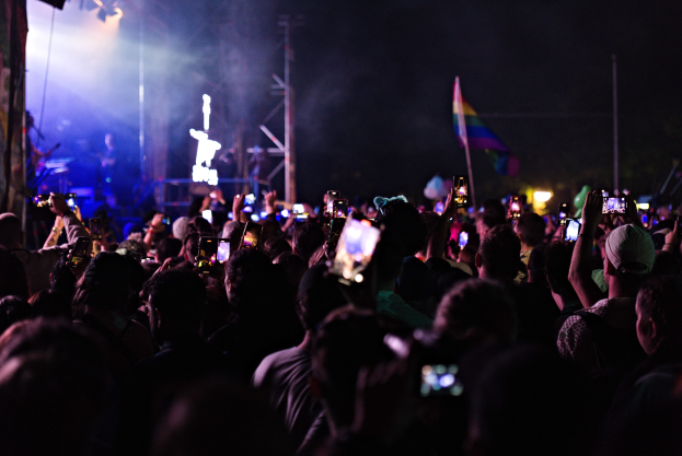 Eine Menge bei einem Konzert hält beleuchtete Handys in die Höhe, während Musiker auf der Bühne spielen und von Lichtmasten und einer Flagge umgeben sind.