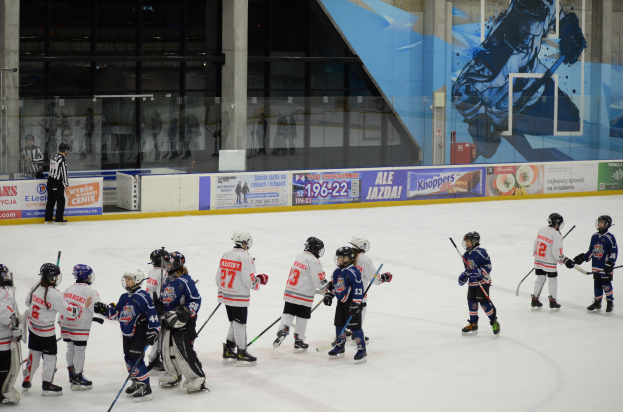 Gruppe junger Menschen beim Eishockey auf einer Indoor-Eisfläche, mit Helmen und Stöcken, einem Zuschauer links und Bannern, Säulen und Glaswänden im Hintergrund.