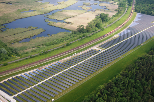 Eine Luftaufnahme eines Solarparks mit zahlreichen Solarmodulen in einem Feld, umgeben von Bäumen, Gras, Wasser und einer nahen Bahntrasse.