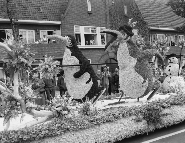 Schwarz-weiß-Foto eines Paradewagens mit Blumen und Pinguin-Figuren, mit Menschen, Bäumen und Gebäuden im Hintergrund.