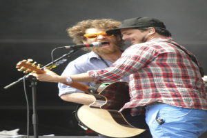 Zwei Männer auf der Bühne, einer in rot-blau kariertem Hemd mit blauen Hosen und der andere mit Goggles und schwarzer Mütze, beide singen in Mikrofone und spielen Gitarren.