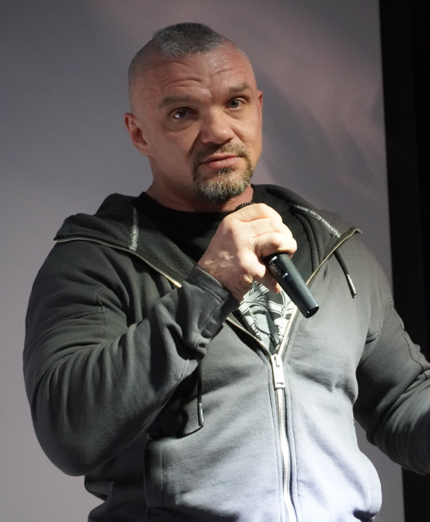 Ein Mann in einer schwarzen Kapuzenjacke, schwarzem Anzug und weißem Hemd, mit zurückgekämmtem Haar, hält ein Mikrofon und steht selbstbewusst vor einer Wand.