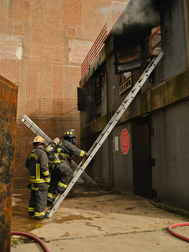 Feuerwehrleute mit Helmen klettern an einer Leiter zu einem rauchgefüllten Gebäude mit Metallobjekten links und Rohren an der Basis.