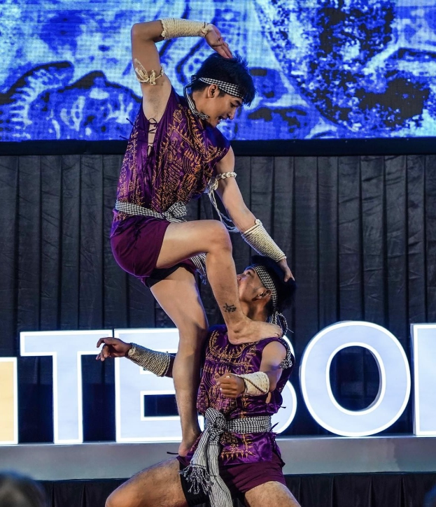 Zwei Performer in violetten Kleidern führen einen akrobatischen Hebemanöver auf der Bühne vor einem Publikum aus, mit einem schwarzen Vorhang und Bildschirm im Hintergrund.
