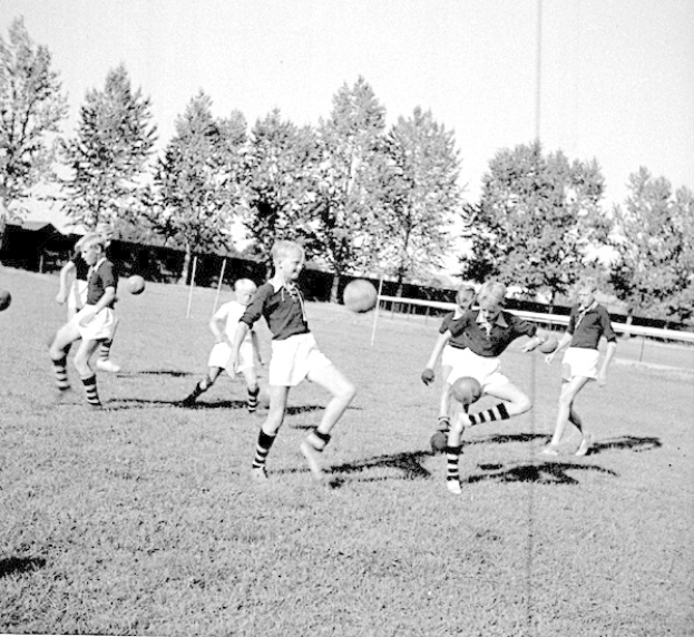 Schwarzes und weißes Foto von jungen Jungs, die auf einem Feld mit Bäumen und einem Pfosten im Hintergrund Fußball spielen.