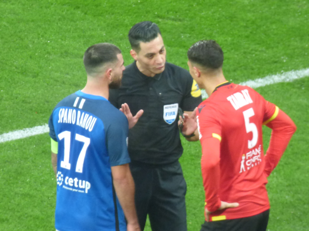 Schiedsrichter spricht mit zwei Männern auf einem Fußballfeld, einer in schwarzer Hemd und Hose, der andere in einem blauen und roten T-Shirt mit Text und Brille.