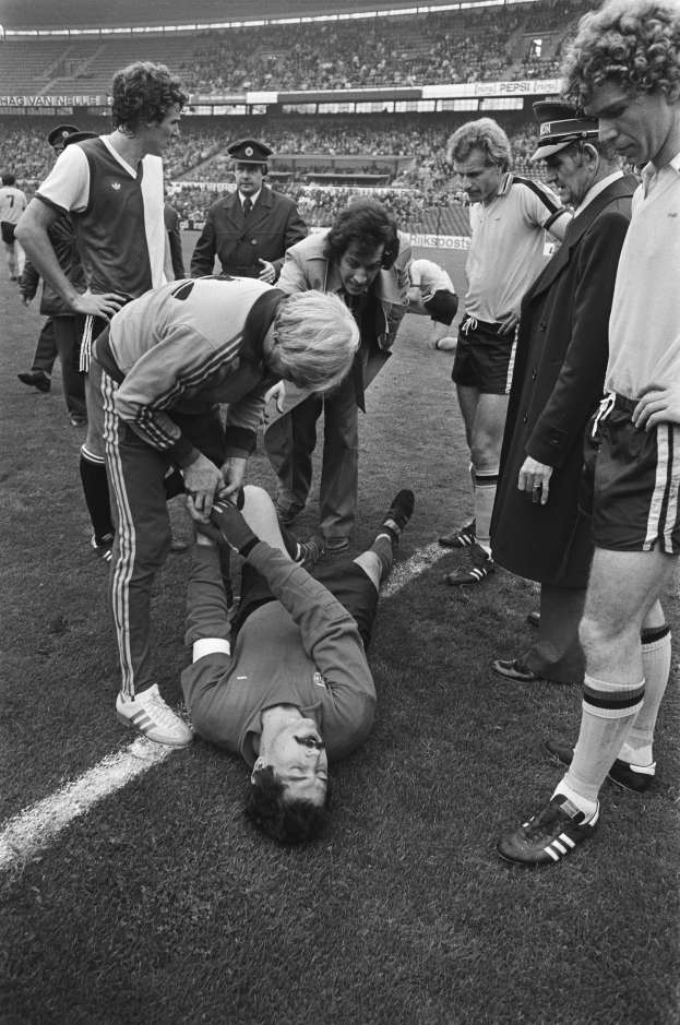 Schwarzes und weißes Bild eines Fußballspielers, der auf dem Boden liegt und von einer Gruppe Männer und einem Stadionpublikum umgeben ist, wobei er verletzt zu sein scheint.