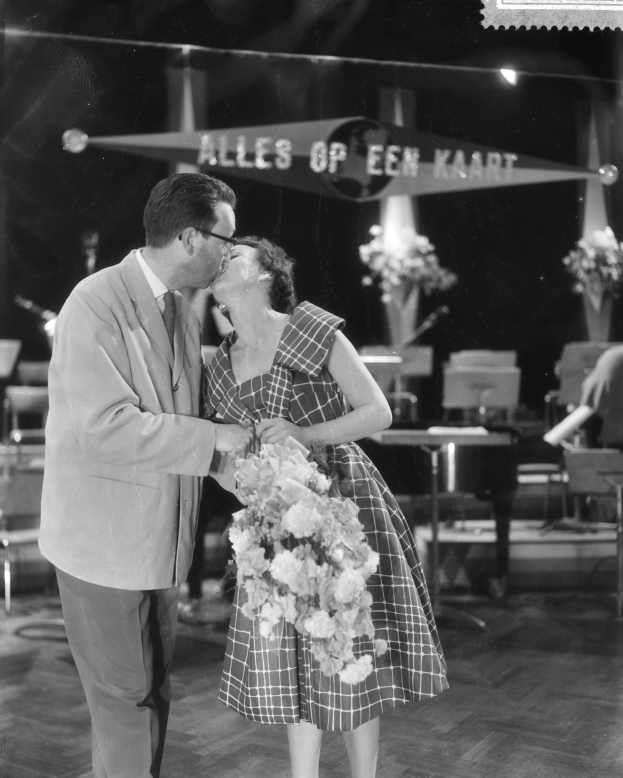 Ein Mann und eine Frau umarmen und küssen sich auf einer Bühne, wobei die Frau einen Blumenstrauß hält und von Stühlen, Blumenvasen und anderen Gegenständen umgeben ist.