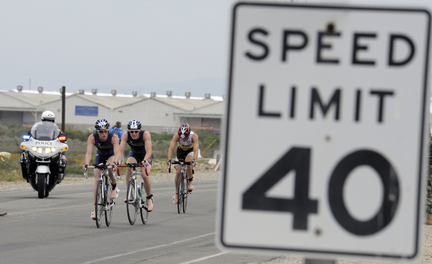 Gruppe von Radfahrern in Helmen auf einer Straße mit einem Geschwindigkeitsbegrenzungsschild, Gebäuden und Bäumen im Hintergrund unter einem klaren blauen Himmel.