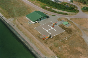 Ein Hubschrauber vor einem Gebäude geparkt, umgeben von Bäumen, Straßen und Fahrzeugen, mit einem Gewässer auf der linken Seite.