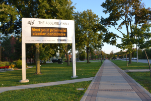 Ein Schild auf dem Boden mit der Aufschrift "Die Versammlungshalle - Treffen Sie Ihre Kandidaten für die Landtagswahl", umgeben von Gras, einem Weg, Bäumen, blühenden Pflanzen, Straßenlaternen, Fahrzeugen, einem Gebäude mit Fenstern und einem bewölkten Himmel.