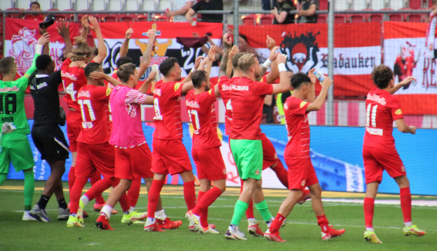 Gruppe von Fußballspielern, die auf einem Feld mit Bannern, einem Zaun und Tribünen im Hintergrund feiern.