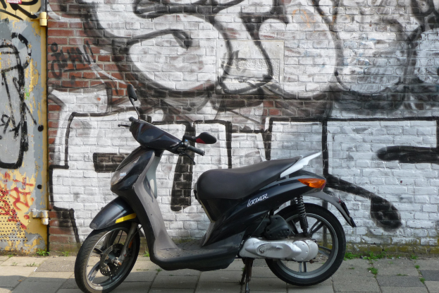 Ein vor einer Backsteinwand mit Graffiti geparktes Motorroller mit einer auf der linken Seite des Bildes sichtbaren Tür.