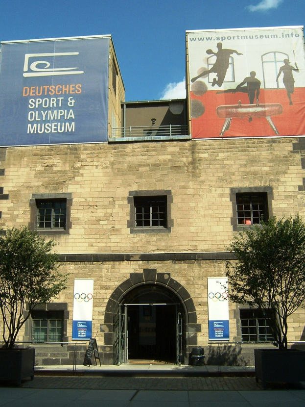 Übersicht des Deutschen Sport & Olympiamuseums in Berlin, mit einem Gebäude mit Fenstern und einer Tür, umgeben von Bäumen und Bannern, unter einem bewölkten Himmel.