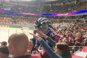 Eine Gruppe von Menschen steht vor einem Publikum bei einem Hockey-Spiel, mit einer Person in einem Kostüm in der Mitte, umgeben von einem Metallzaun, einem Banner mit Text, Metallstangen und einer Decke mit Deckenleuchten.