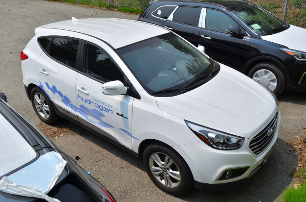 Drei wasserstoffbetriebene Autos auf einem Parkplatz mit Maschendrahtzaun im Hintergrund und Gras auf der rechten Seite geparkt.