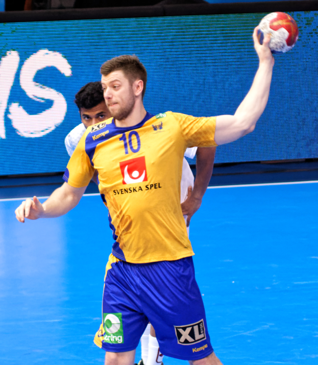 Ein Mann in einem gelben T-Shirt und blauen Shorts hält einen Handball auf einem Feld, mit einem Hintergrundbild, das "Svenský Spel Handball World Cup 2019" anzeigt.