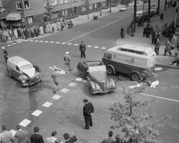 Eine Schwarz-Weiß-Szene eines Autounfalls an einer Straßenkreuzung mit mehreren Fahrzeugen, darunter ein Lieferwagen, und einer Gruppe von Menschen, die herumsteht, mit Gebäuden, Bäumen, Pfählen und Texttafeln im Hintergrund.