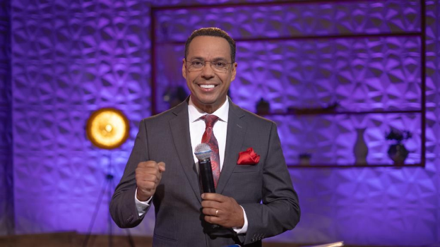 Stephen Colbert, Moderator von "The Daily Show with Trevor Noah", steht auf einer Buhne und halt einen Mikrofon, tragt einen Anzug, Krawatte und Brille, mit einem Licht und Stander hinter ihm.