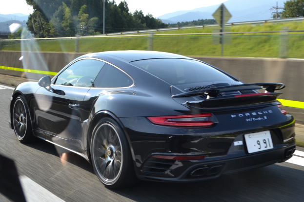 Ein schwarzer Porsche 911 GTS fährt auf einer Autobahn, umgeben von einem Zaun, Gras, Bäumen, Masten, Drähten und Bergen unter einem klaren blauen Himmel.