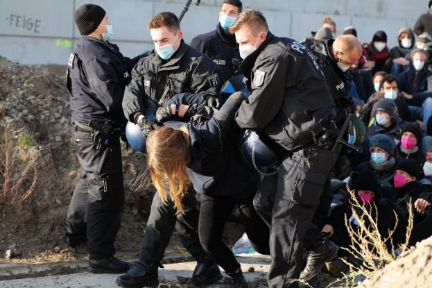 Gruppe maskierter Personen in Mützen, einige bewaffnet, die eine Frau umgeben, die von der Polizei eskortiert wird, mit urbanen Elementen und protestbezogener Graffiti im Hintergrund.