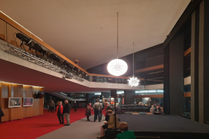 Eine Gruppe von Menschen in einem großen Raum mit rotem Teppich, Foto Rahmen an der linken Wand, Deckenleuchten, einem Geländer und einem Kronleuchter, was die Eröffnungszeremonie des Nationalen Museums der Niederlande anzeigt.