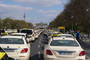 Eine lange Reihe von Taxis parkt am Straßenrand einer belebten Straße in Berlin, Deutschland, mit Fahrzeugen, Radfahrern und Fußgängern, flankiert von Laternenpfählen, Bäumen und Gebäuden, einschließlich eines Bogens und eines Turms.