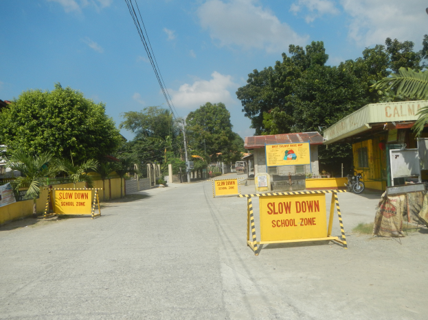 Eine Straße mit einem gelben "Vorsicht Schulbereich"-Schild, parkende Fahrzeuge auf der rechten Seite und im Hintergrund Bäume, Gebäude, Mäste, Drähte und Himmel.