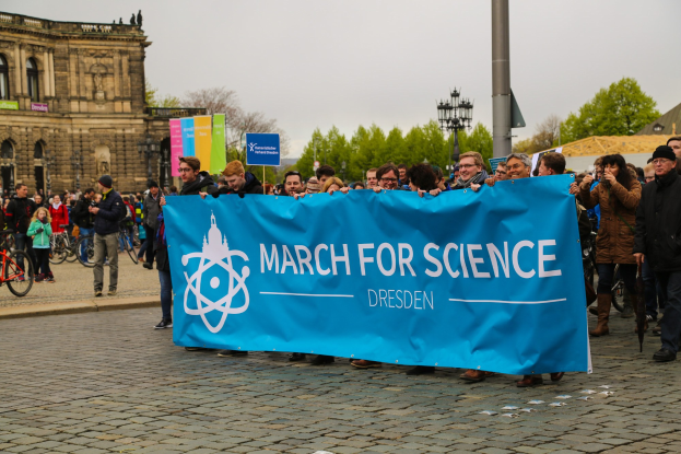 Gruppe von Menschen mit einer "March for Science Dresden"-Plakette vor einem Gebäude mit Fenstern, umgeben von Bäumen, Laternenmasten und Fahrrädern unter einem sichtbaren Himmel.