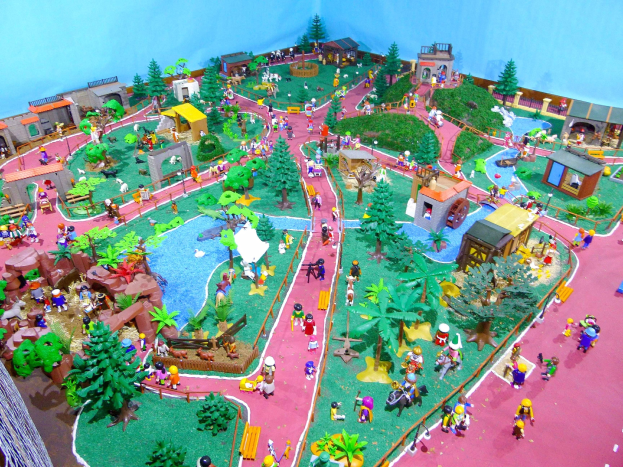 Ein Miniaturmodell eines Zoos mit Besuchern, umgeben von Bäumen, Häusern, Pflanzen, Gras und einem Zaun, vor einer Hintergrundwand mit Text unten.