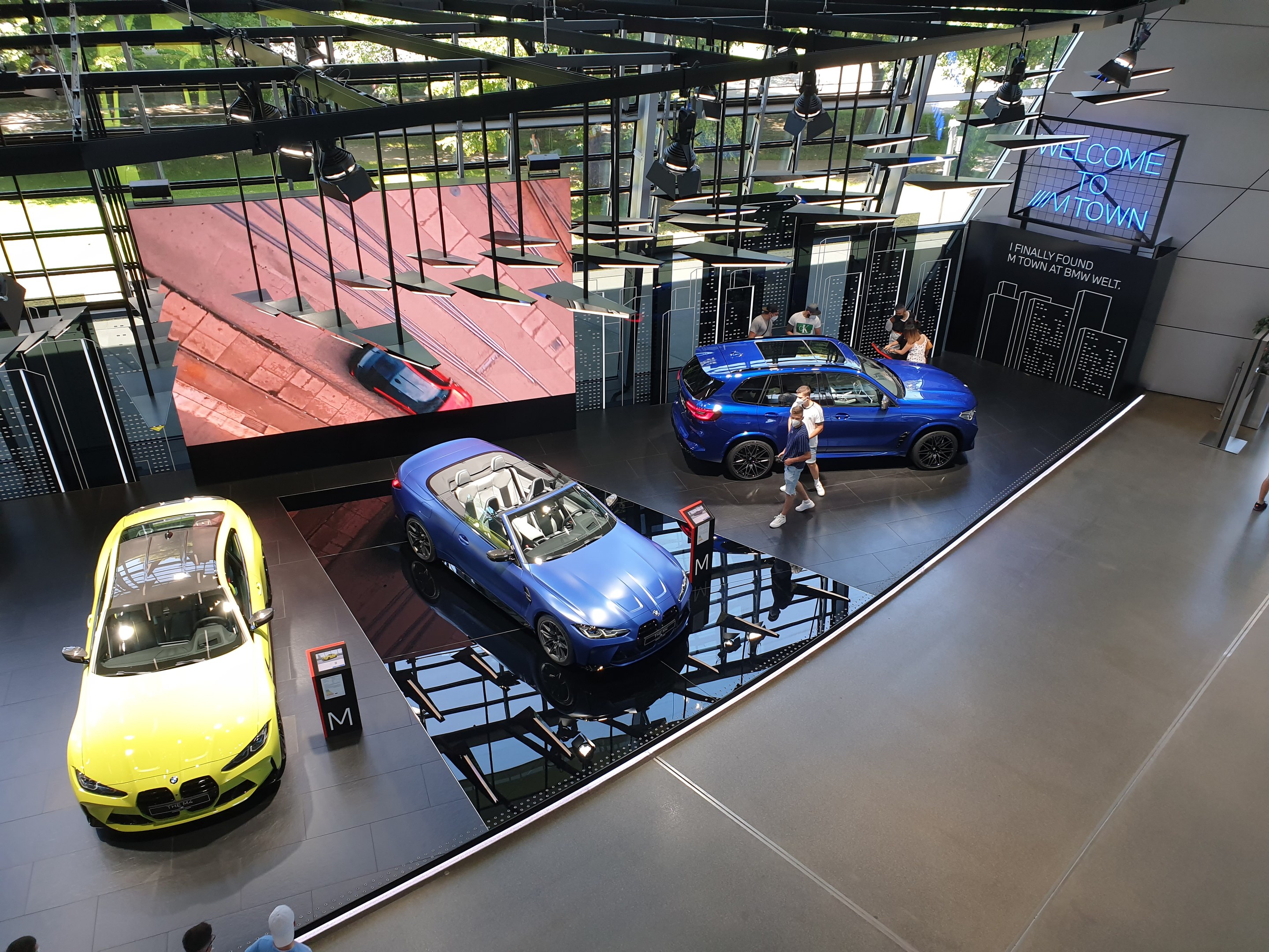 Eine Gruppe von Menschen steht im BMW Museum in München, mit ausgestellt Autos auf der linken Seite und einem Schild mit der Aufschrift "BMW Museum München" auf der rechten Seite, mit Blick auf Bäume, Gebäude und den Himmel durch Glasfenster.