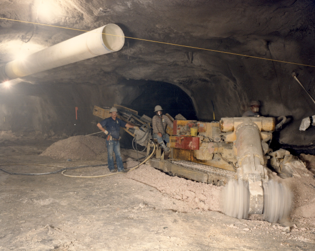 Gruppe von Männern in Helmen, die in einem dunklen Stollensystem mit Maschinen und Rohren arbeiten.
